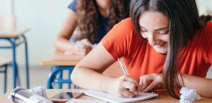 Coesão e coerência: fotografia de uma menina sorrindo enquanto escreve em um caderno em uma sala de aula.