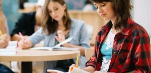 Dicas para o Enem: fotografia de uma sala de aula com estudantes. À frente e em destaque está uma estudante olhando para um livro em seu colo e segurando uma caneta.