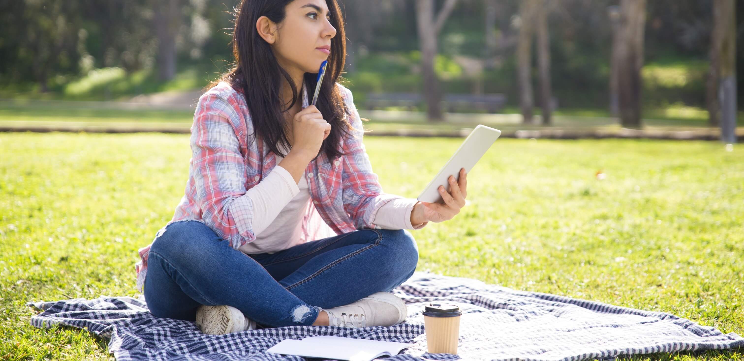 modelos de introdução para redação: imagem de uma mulher sentada no chão em um parque. Ela está segurando um caderno e olhando para o alto com a expressão pensativa.