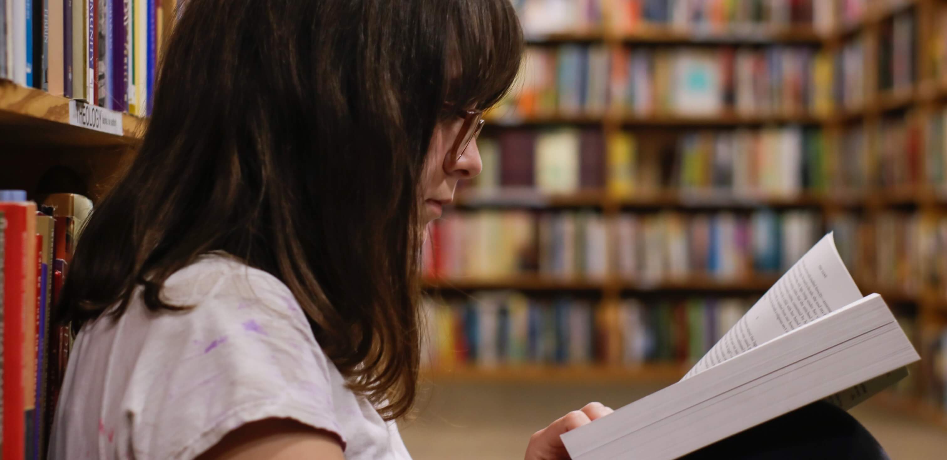 Diferença entre texto expositivo e argumentativo: fotografia de uma menina de óculos sentada no chão de uma biblioteca lendo um livro.