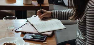 Conectivos para redação - um mulher sentada, escrevendo em seu caderno com um notebook, celular, copo e prato em cima da mesa.