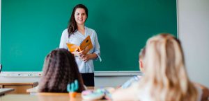 O que é regência verbal e nominal - fotografia de uma sala de aula. À frente do quadro negro e de frente para a foto está a professora com um livro na mão. De frente para a professora estão três alunos sentados nas carteiras.