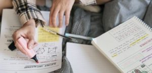 Fotografia de uma pessoa de pernas cruzadas marcando uma folha de estudos. Em cima do seu colo estão livros de estudo e canetas.