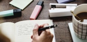 Como melhorar o vocabulário - mesa com marcadores de texto, xícara de café, grampeador, post-it e um caderno sendo escrito por uma pessoa.
