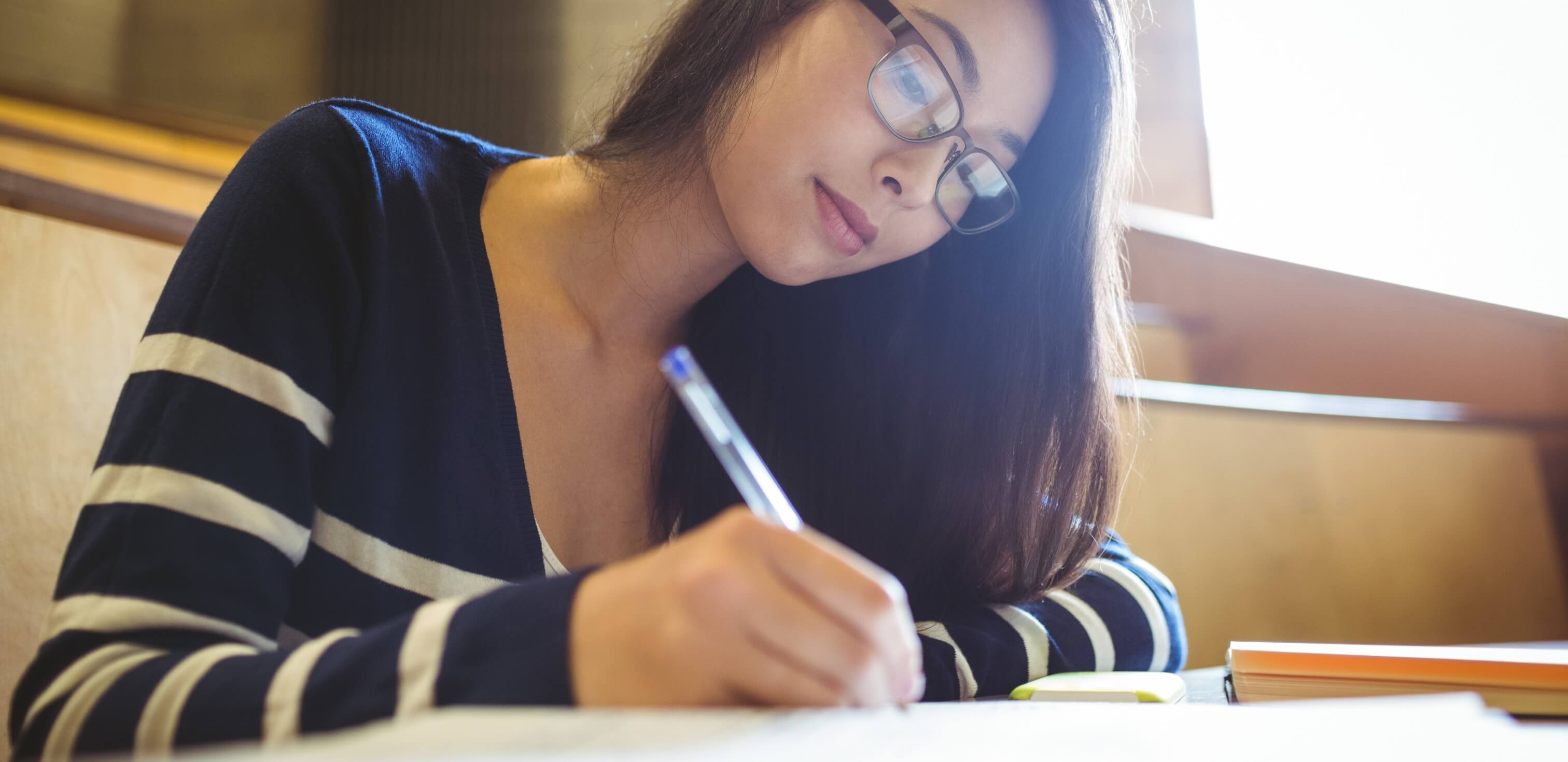 Como estudar redação: fotografia de uma menina de óculos escrevendo em um caderno.
