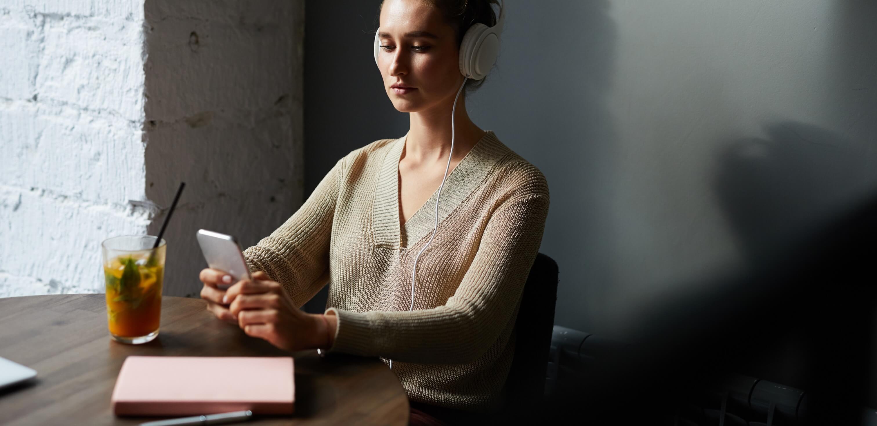 Fotografia de uma mulher sentada à mesa ouvindo podcasts para estudar com fones de ouvido em seu celular.