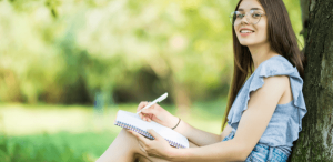Eixos temáticos: imagem de uma menina escrevendo em seu caderno sentada em um parque com muitas árvores
